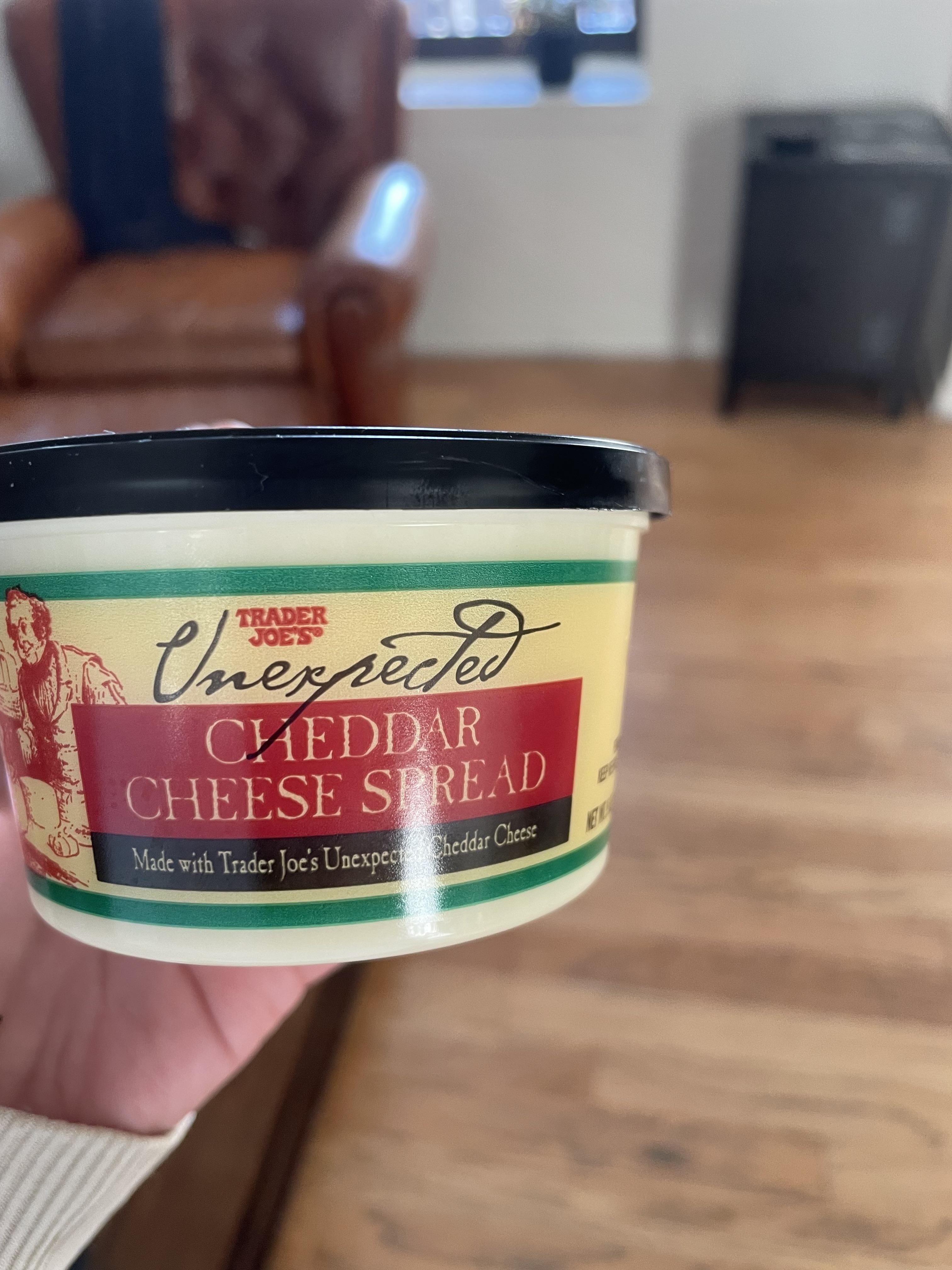 A hand holding a container of Trader Joe's Unexpected Cheddar Cheese Spread. Background features a wooden floor, a leather chair, and a black cabinet