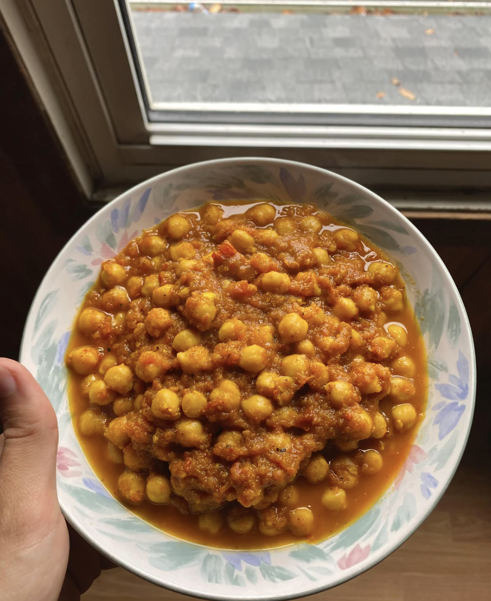 A close-up of a hand holding a bowl of chickpea curry near a window. The curry is thick with chickpeas in a spiced tomato-based sauce