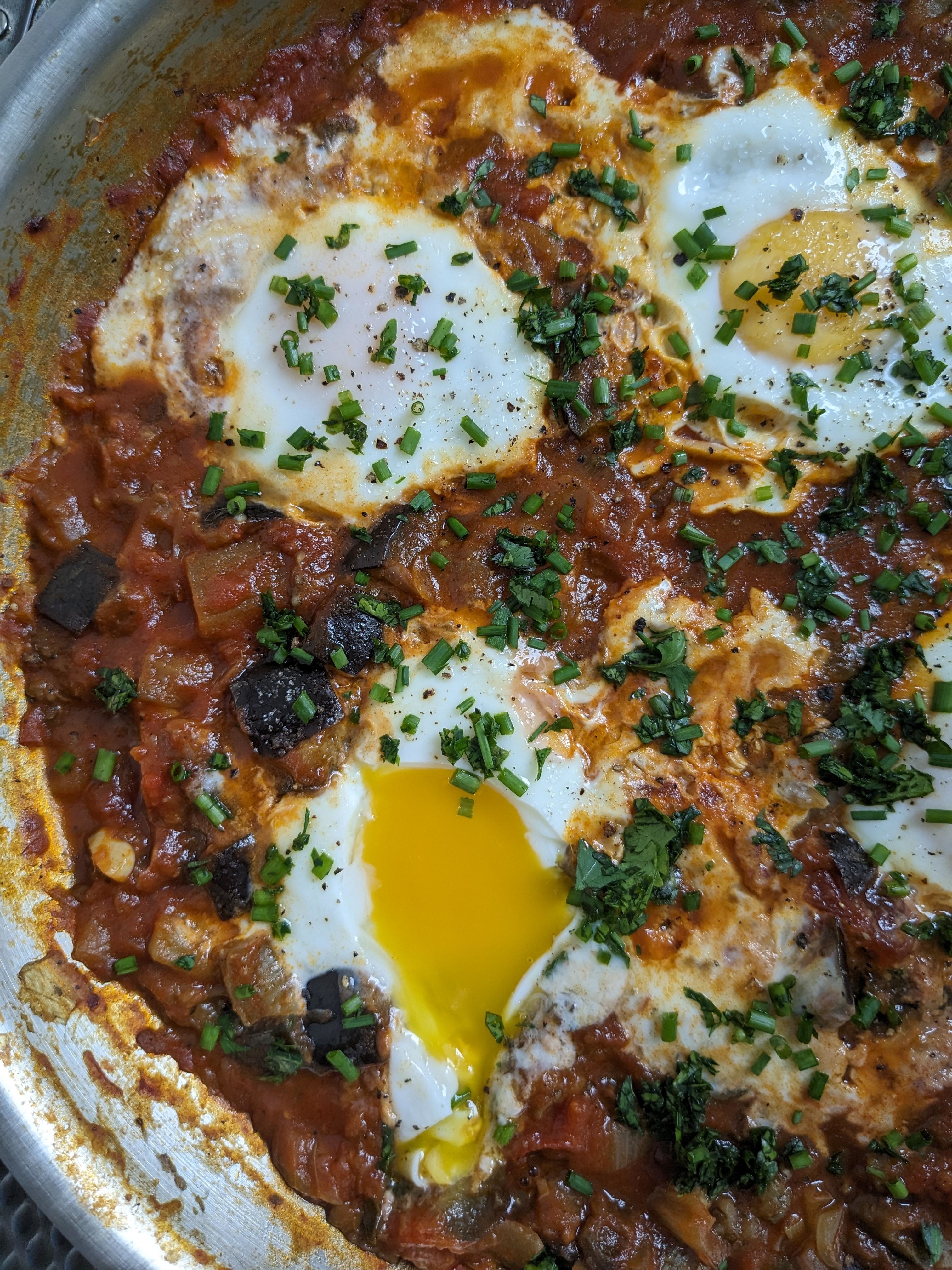 A dish of shakshuka with poached eggs in a tomato and vegetable sauce, garnished with chopped herbs. One egg yolk is broken and partially oozing