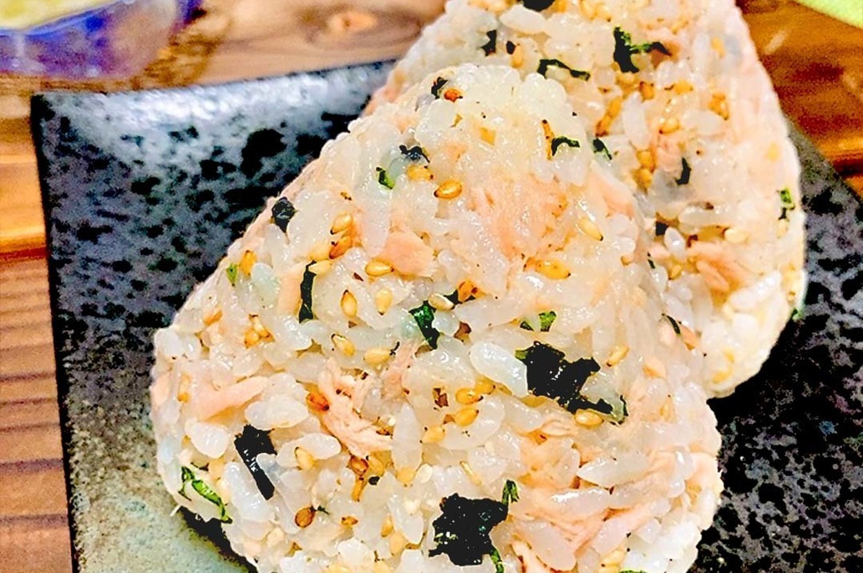 おにぎりが二つ、黒いお皿の上に置かれている。背景には別の皿やお茶碗が見える。