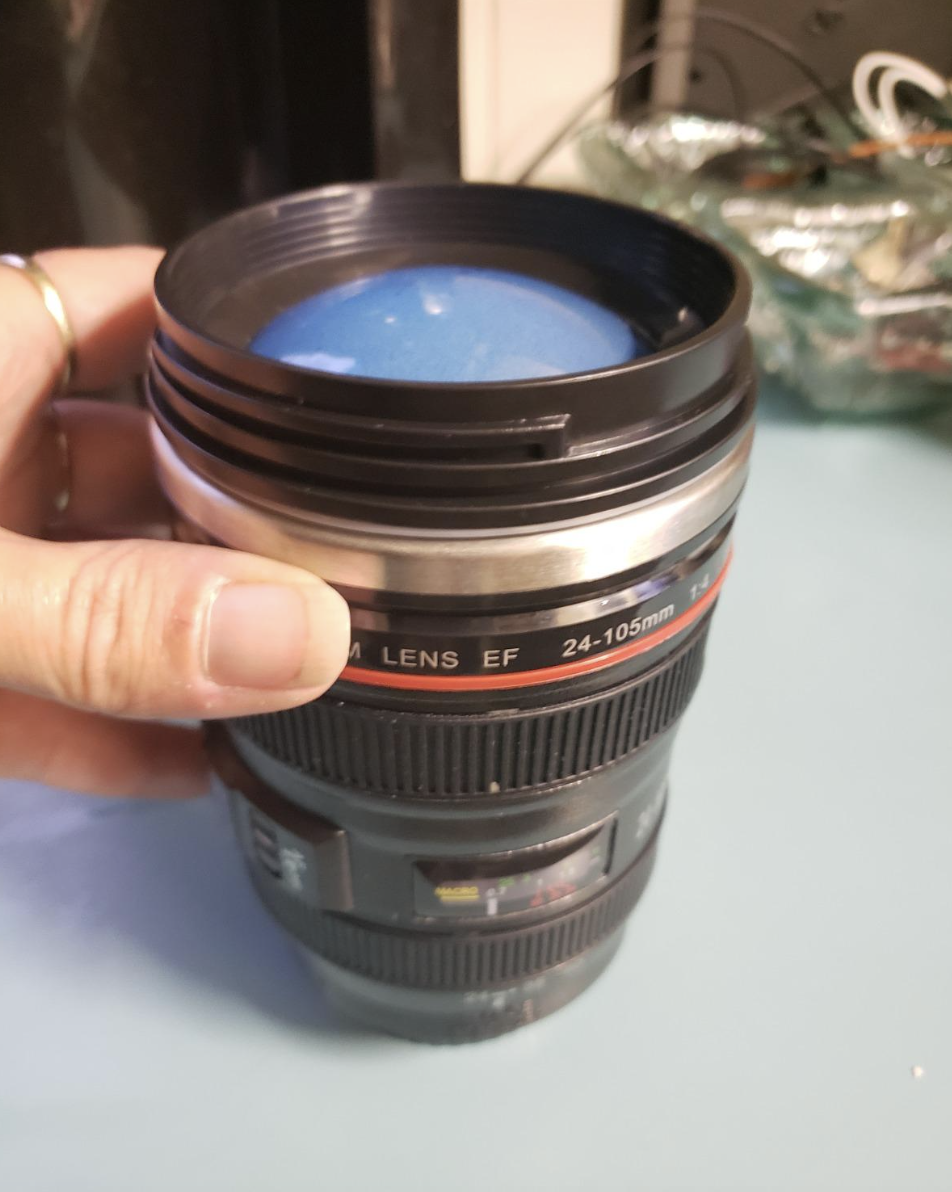 A hand holds a novelty camera lens mug, labeled 