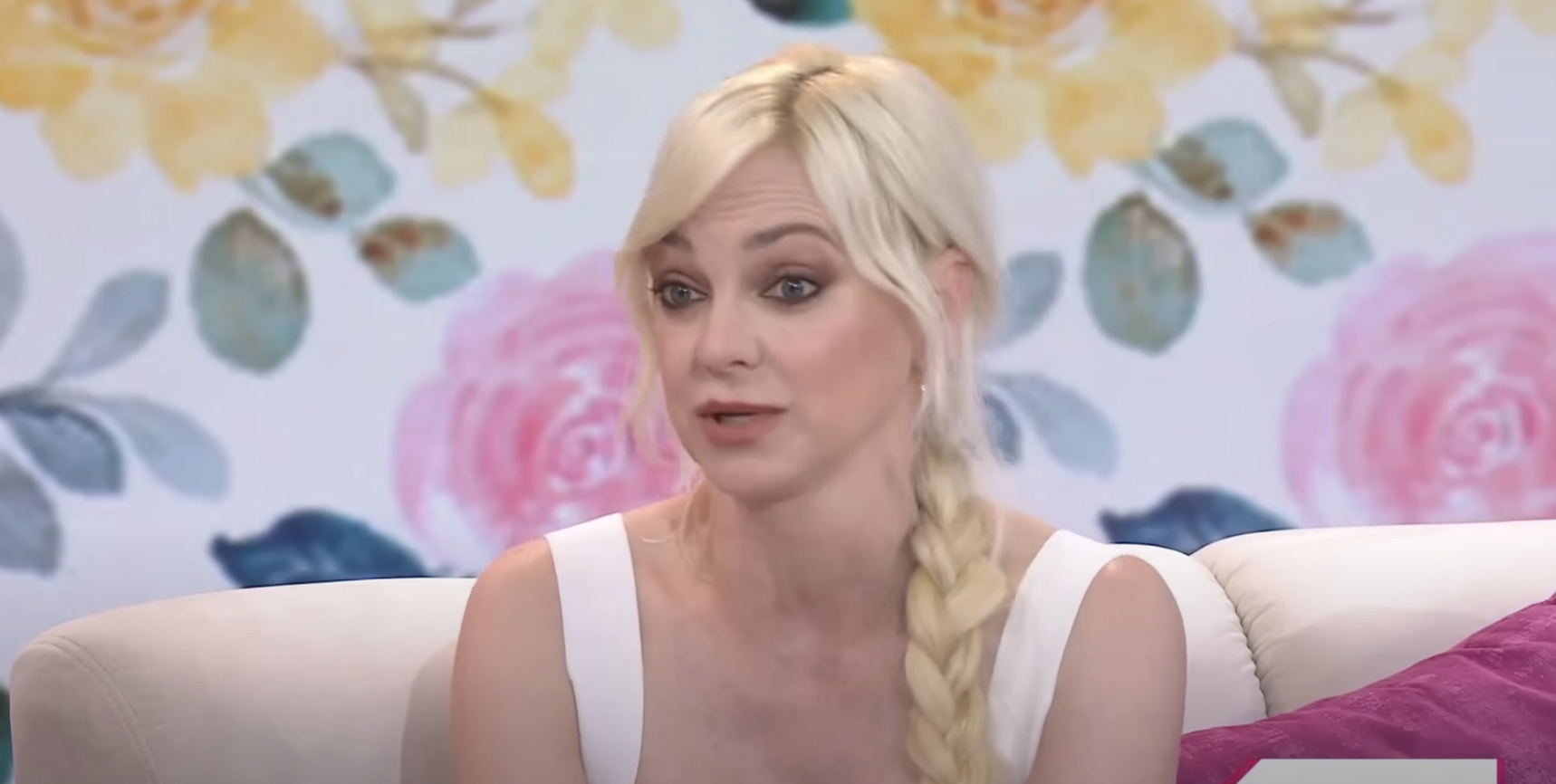 Anna Faris, in a white dress with a braided hairstyle, speaks during an interview against a floral background