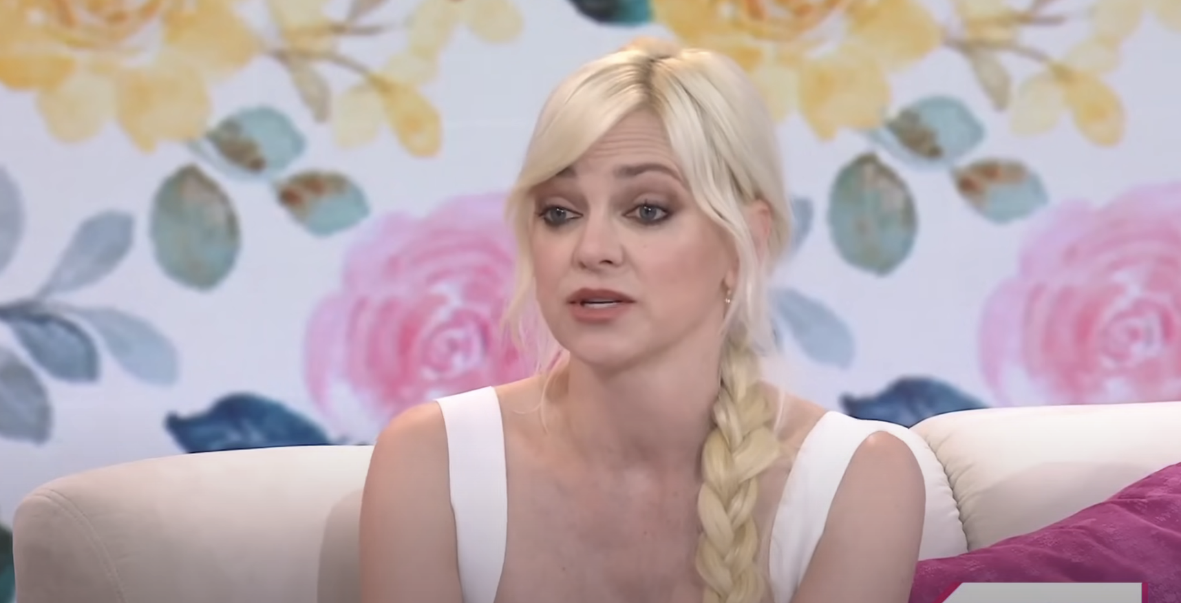 Anna Faris in a white sleeveless dress with a long braid, sitting on a couch with a floral background