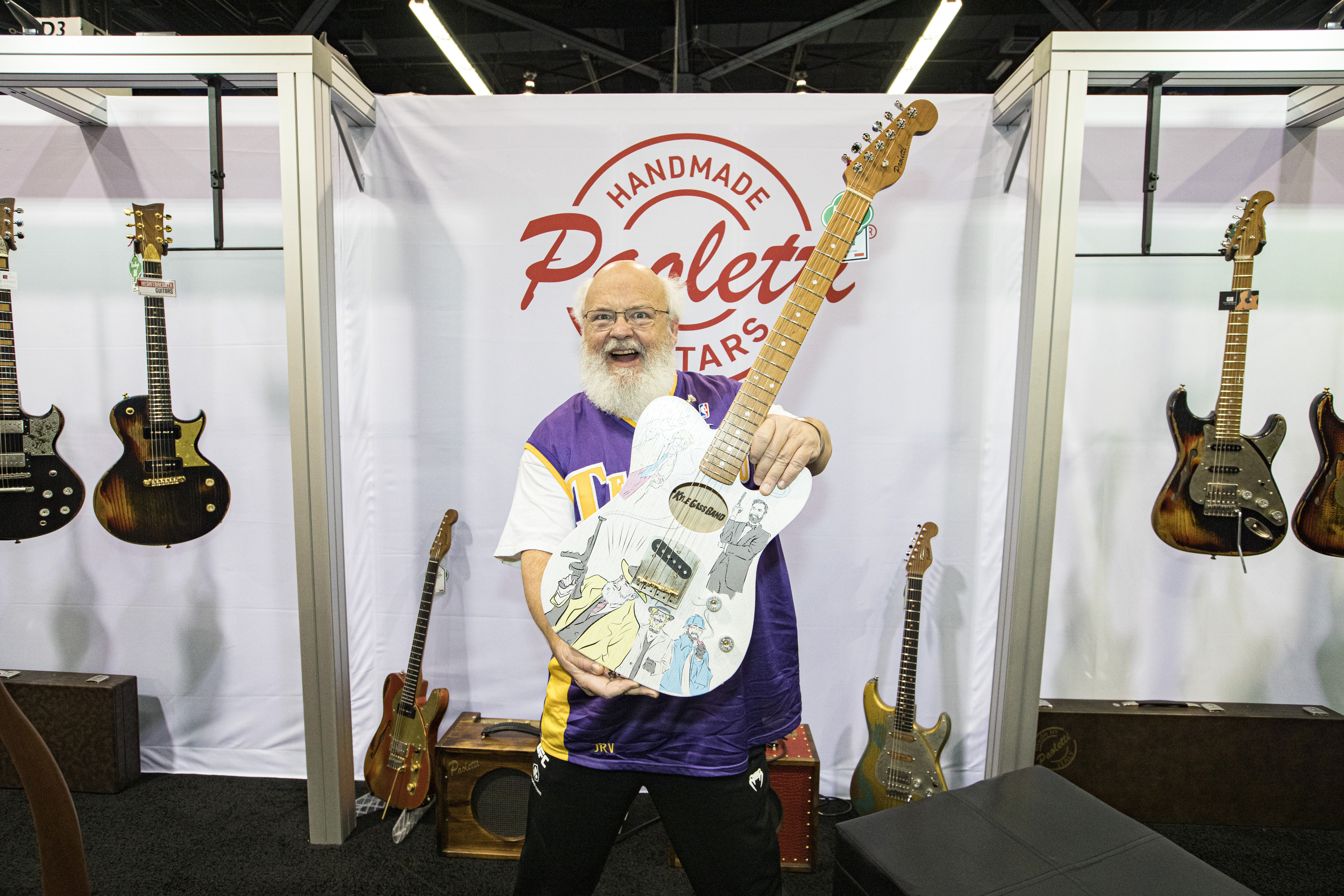 A bearded man, wearing a sports jersey, happily holds up a custom-designed electric guitar in front of a "Handmade Rosetta Guitars" banner at a music trade show