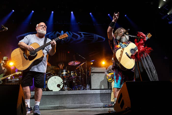 Jack Black and Kyle Gass performing onstage with guitars as part of Tenacious D. Jack Black is wearing vibrant, colorful attire with a feathered glove