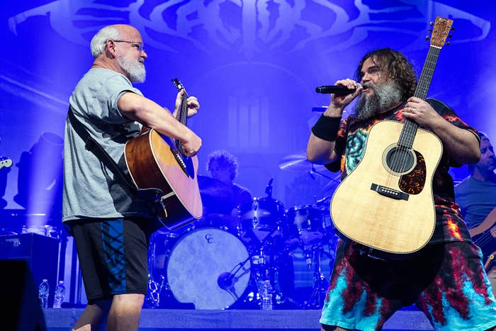 Jack Black and Kyle Gass of Tenacious D perform on stage, playing guitars and singing passionately, with a drummer visible in the background