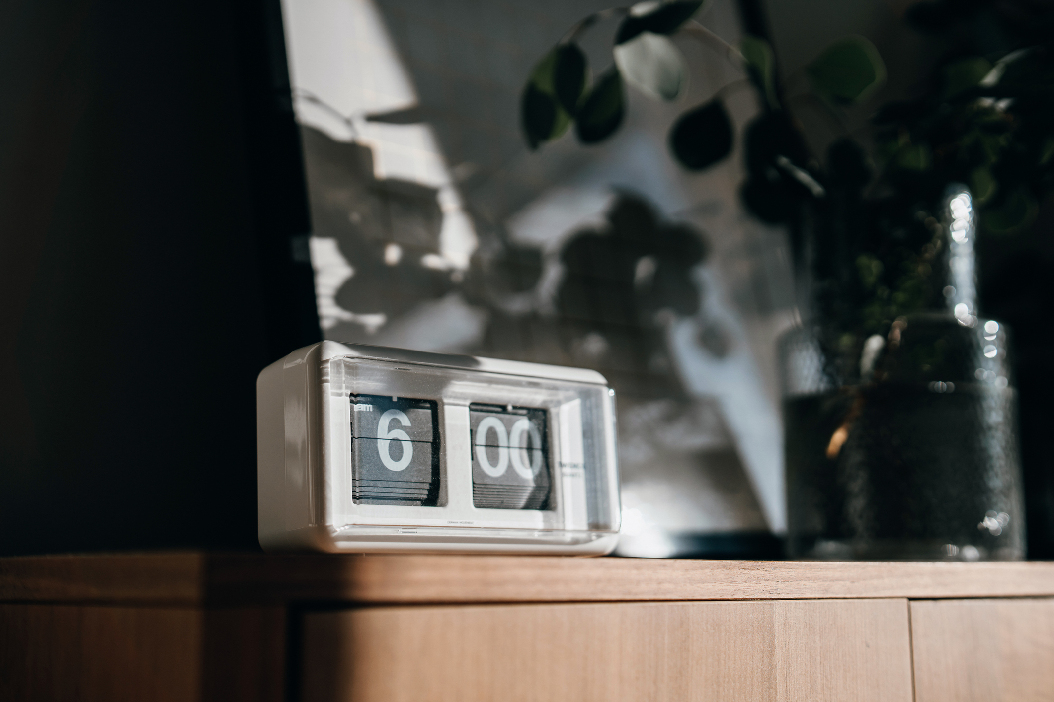 A retro flip clock displaying 6:00 sits on a wooden cabinet with a plant in the background