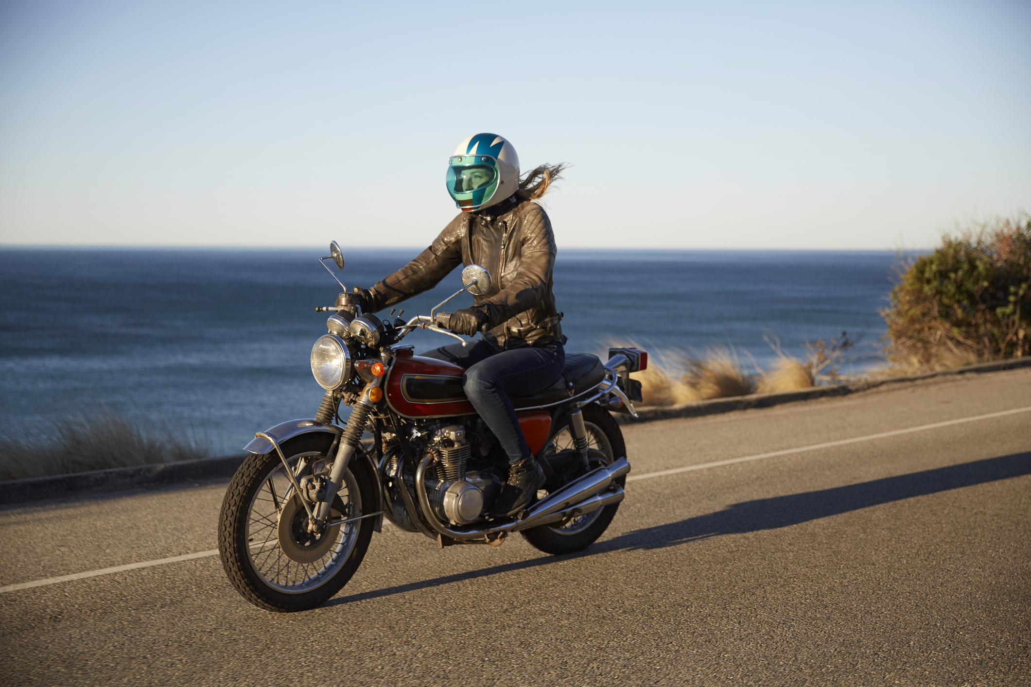 Person riding a motorcycle along a coastal road with the ocean in the background, wearing a helmet and leather jacket