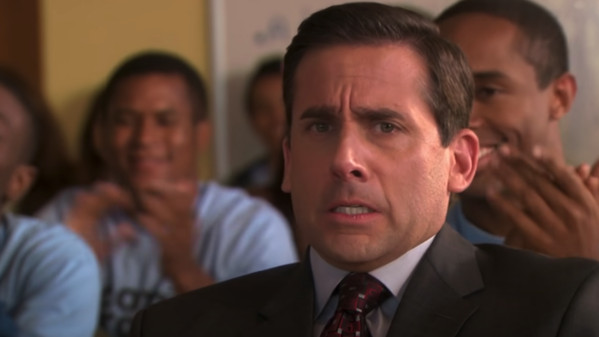 Steve Carell in a suit and tie, looking concerned. People in the background are clapping and seemingly happy