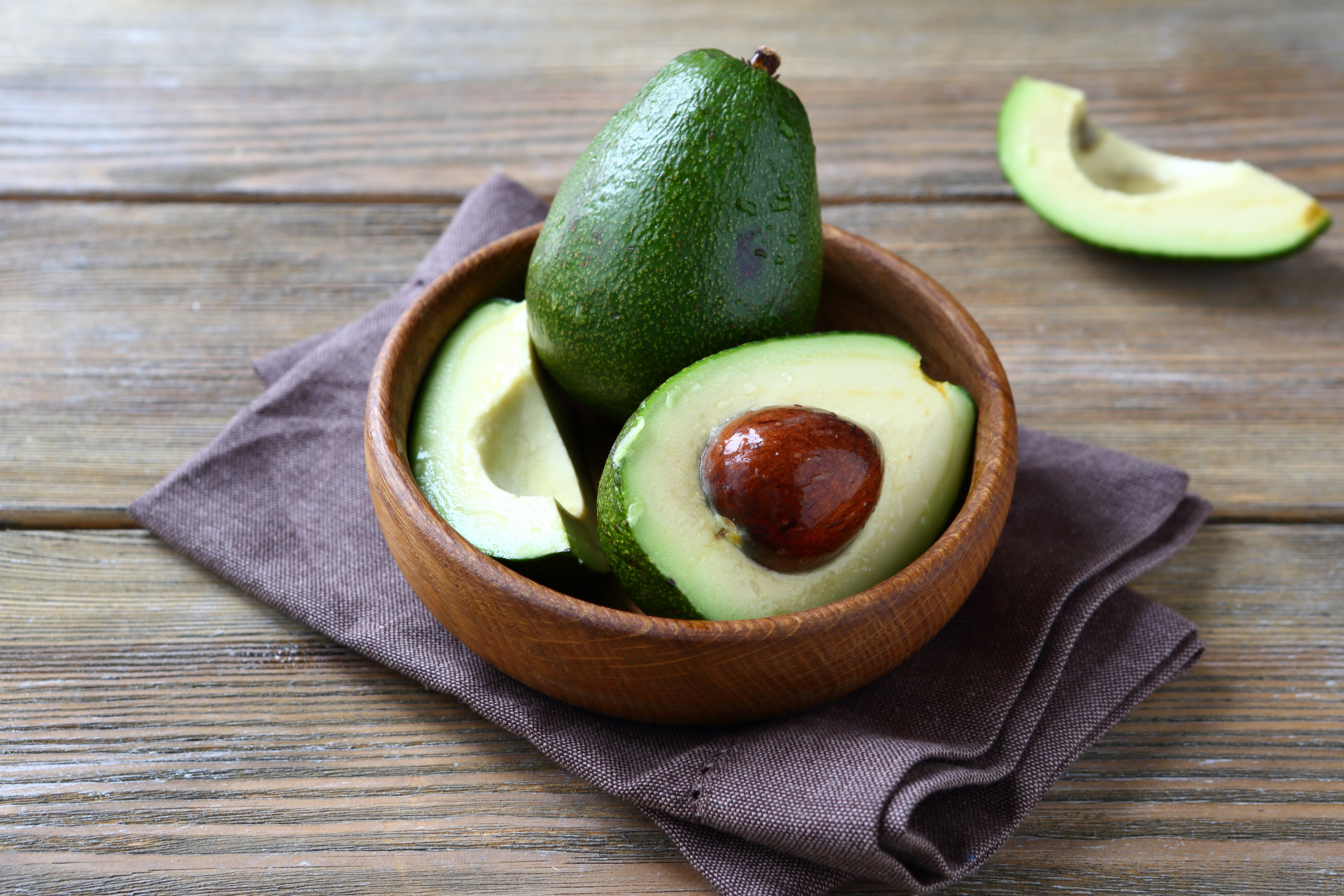 A wooden bowl with whole and sliced avocados on a cloth napkin placed on a wooden surface