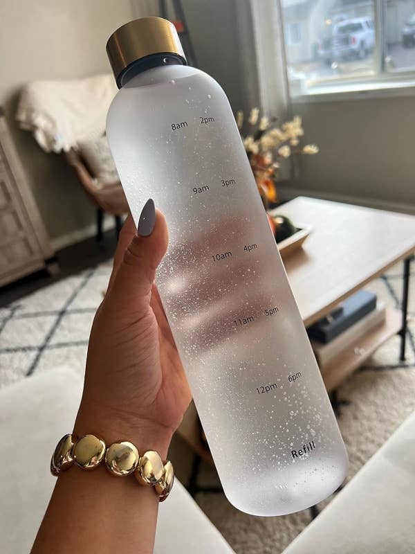 Hand wearing a gold bracelet holds a frosted water bottle with time markers from 8am to 6pm and a reminder to refill, promoting regular hydration