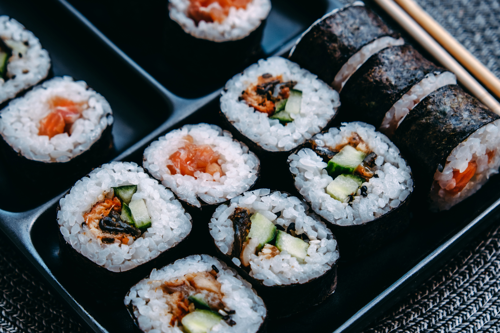 Close-up of a sushi tray featuring several pieces of sushi rolls with various fillings, including cucumber, salmon, and crunchy toppings, next to chopsticks