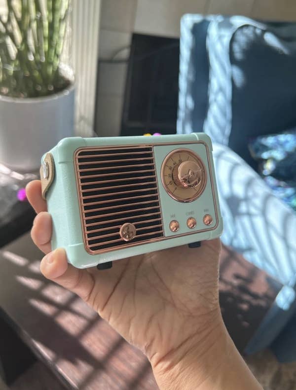 A hand holding a small retro-styled portable radio with a circular dial and buttons. A potted plant and furniture are in the background