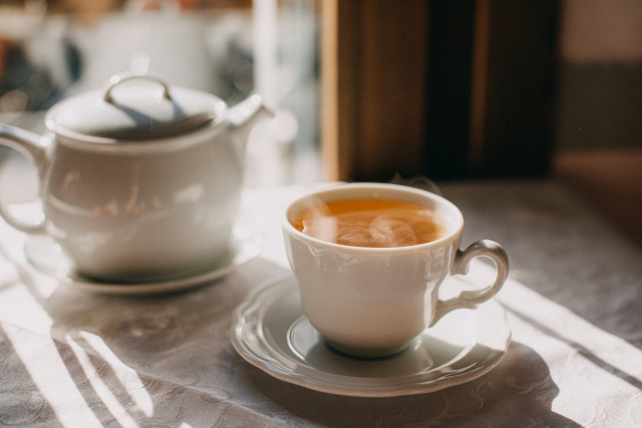 A white tea cup filled with hot tea sits on a matching saucer next to a white teapot on a sunlit table by a window