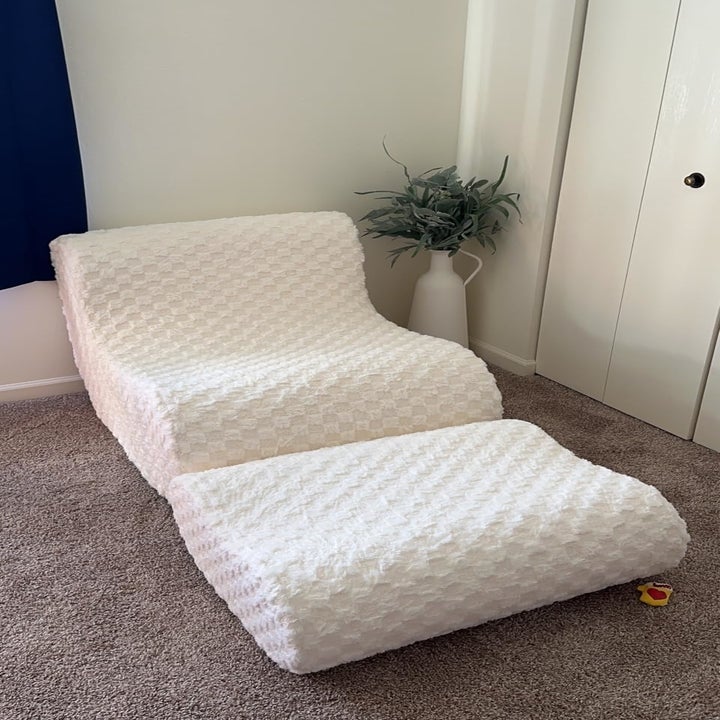 A white, textured lounge chair with an ottoman is placed on a carpeted floor next to a white vase with greenery