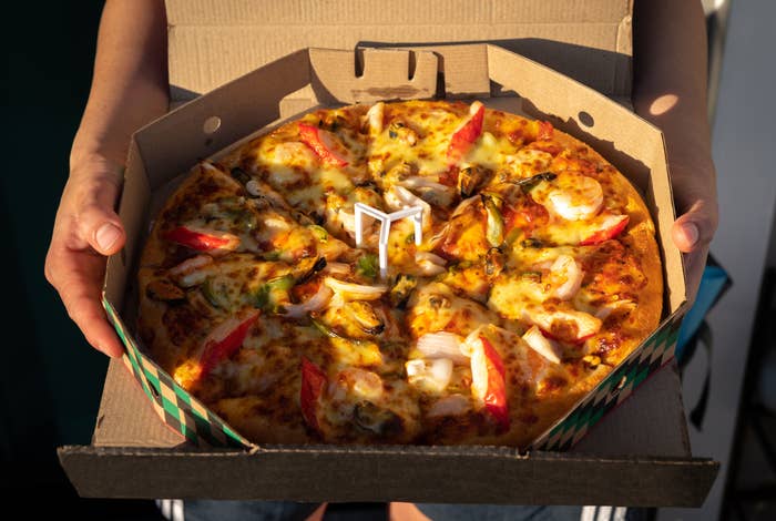 Person holding a pizza with shrimp, onions, and red bell pepper slices in a cardboard box