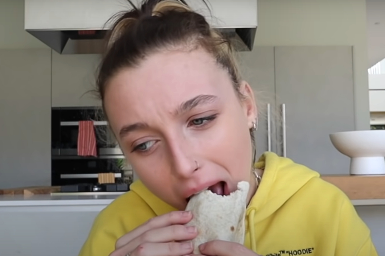 Emma Chamberlain takes a bite of a burrito while seated in a kitchen wearing a hoodie