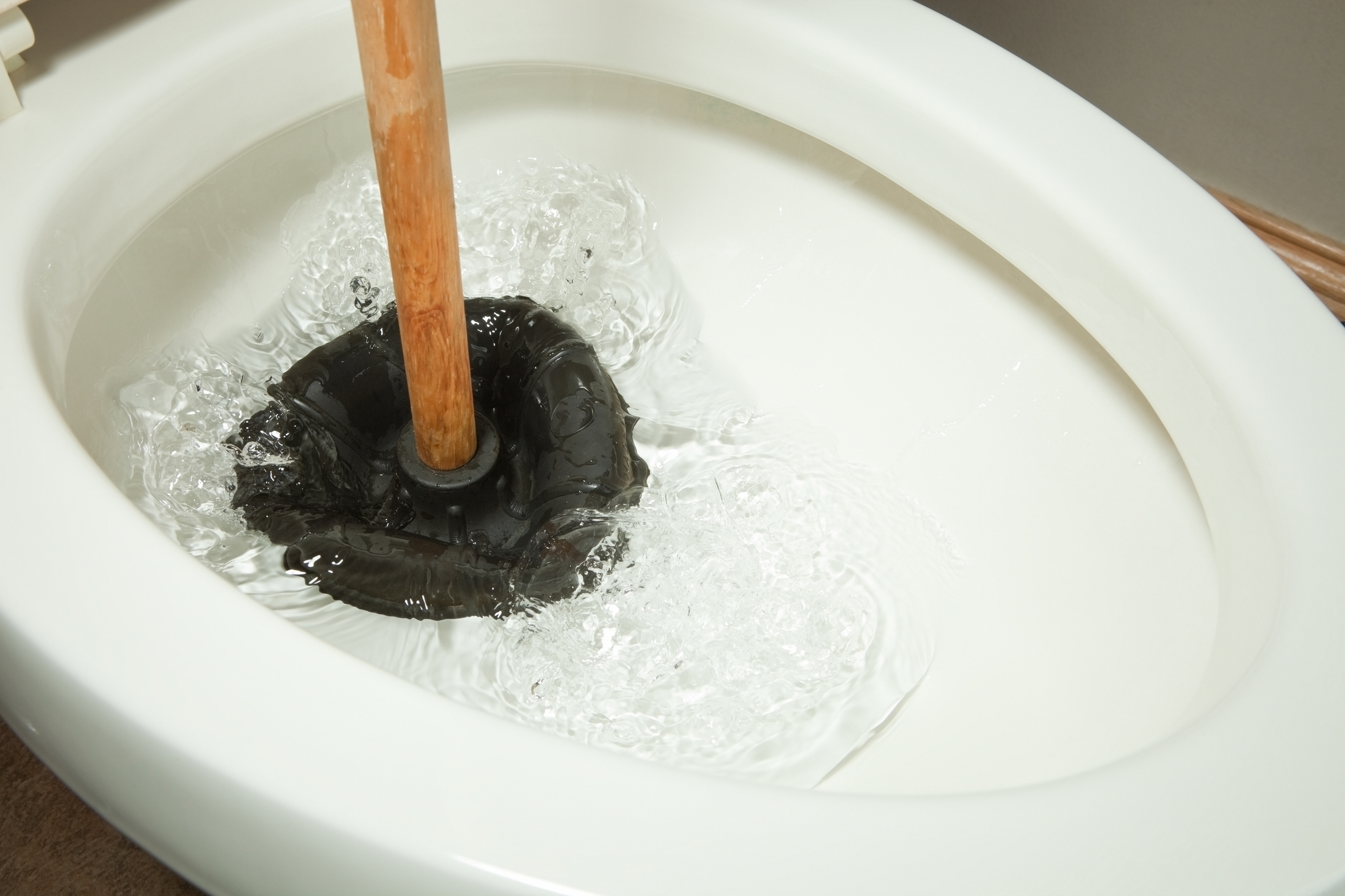 A toilet bowl being cleaned with a wooden-handled plunger. Water is splashing around the plunger