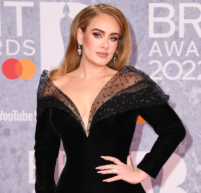 Adele at the BRIT Awards 2022, wearing a gown with a deep V-neckline and sheer, polka-dotted sleeves. She is posing with one hand on her hip