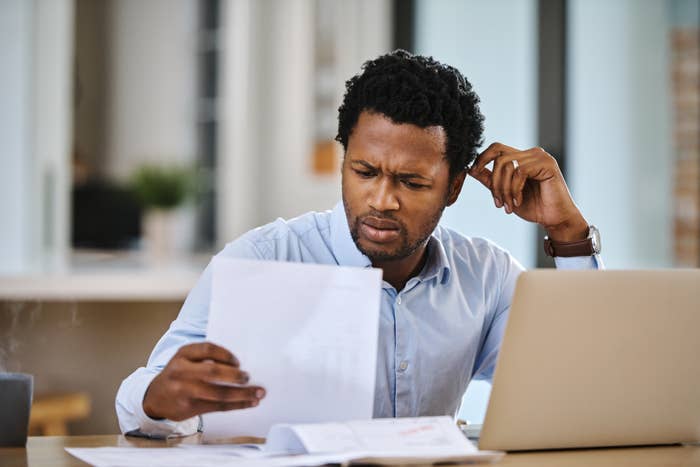 A person in a light shirt sits at a desk, looking concerned while holding a document. A laptop and papers are on the desk