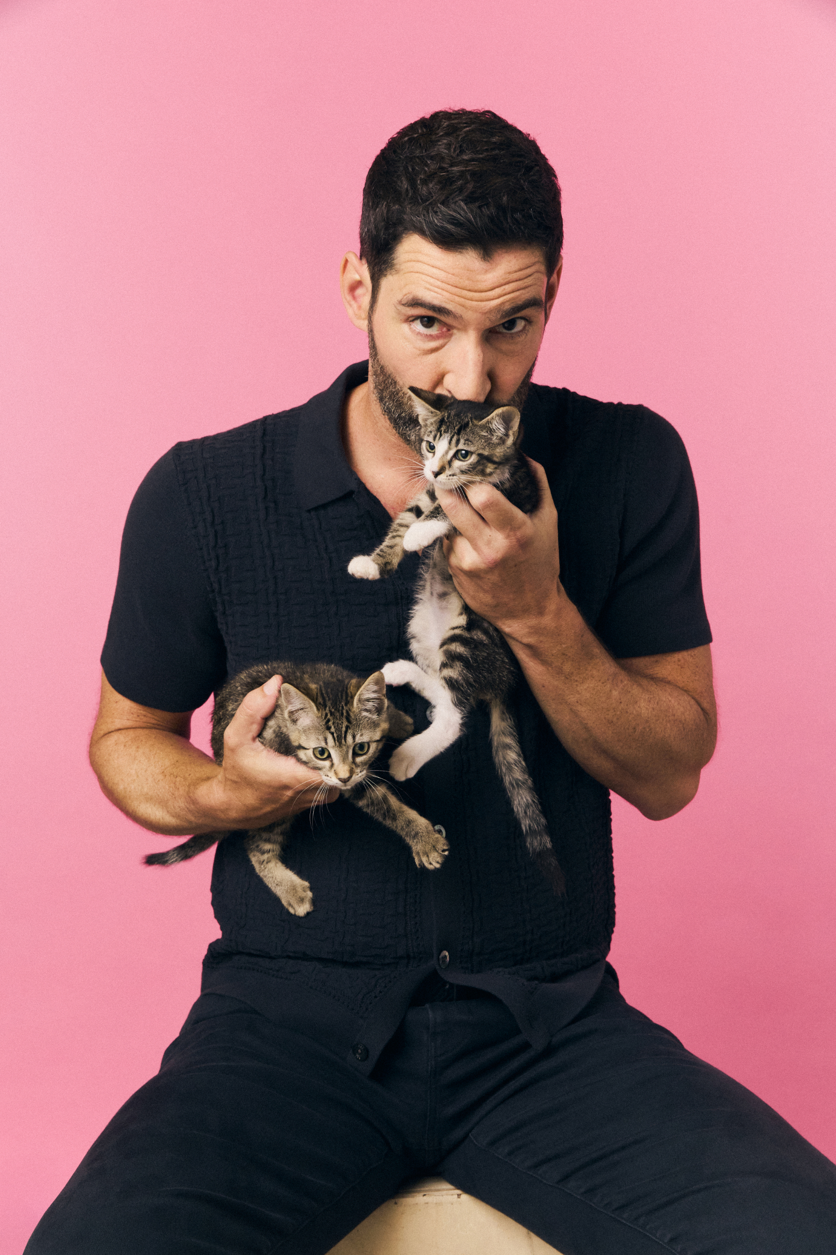 Tom Ellis seated on stool, wearing a black shirt and pants, holding two kittens against a pink backdrop