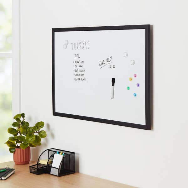 Whiteboard on wall lists &quot;Tuesday To-Do: 1. Make App, 2. Do Laundry, 3. Plan Budgets, 4. Clean Kitchen, 5. Water Plants&quot;; includes notes on a bake sale and a marker. Desk with plant below