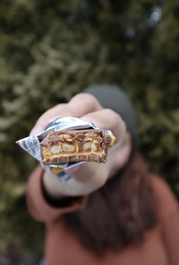 A person is holding an open candy bar with visible nuts and caramel. The focus is on the candy bar, with the person’s face blurred in the background