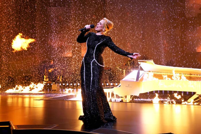 Adele performs on stage in a\ sequined gown, holding a microphone, with flames and a white grand piano in the background
