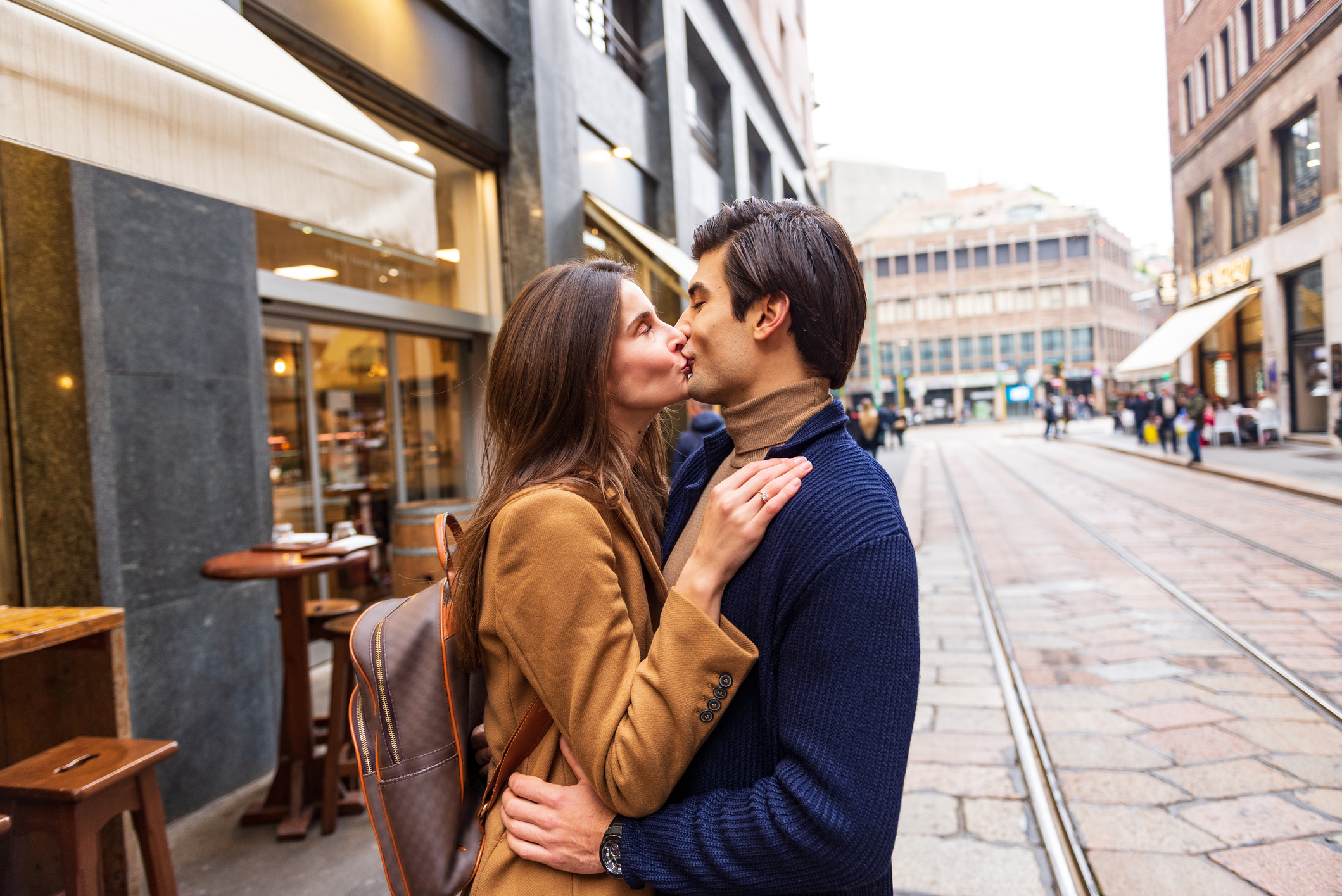A couple kisses on a busy city street with people walking in the background. The woman wears a coat and backpack, while the man wears a sweater
