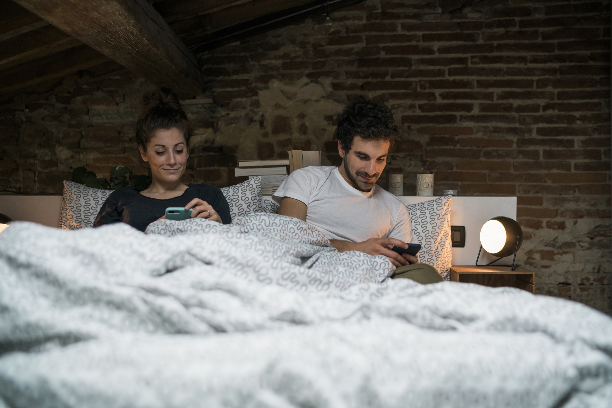 A man and woman are sitting in bed under the covers, both using their smartphones. The room has exposed brick walls and soft lighting