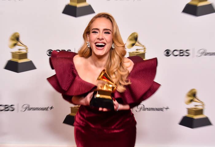 Adele, smiling and holding a Grammy award, wearing an elegant dress with large ruffle sleeves at a red carpet event