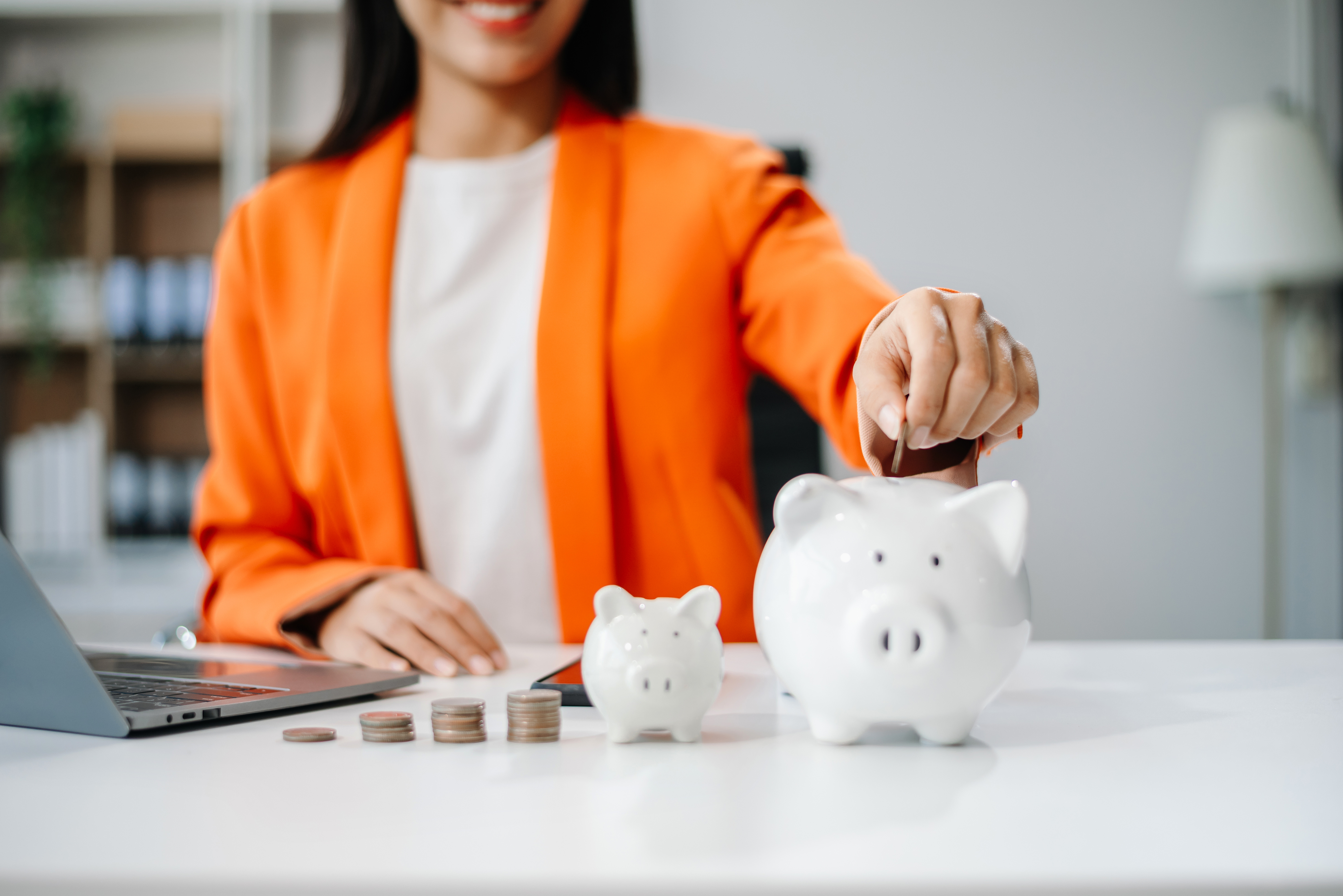 A person in a blazer places a coin into a small piggy bank beside a larger one at a desk with a laptop and stacked coins