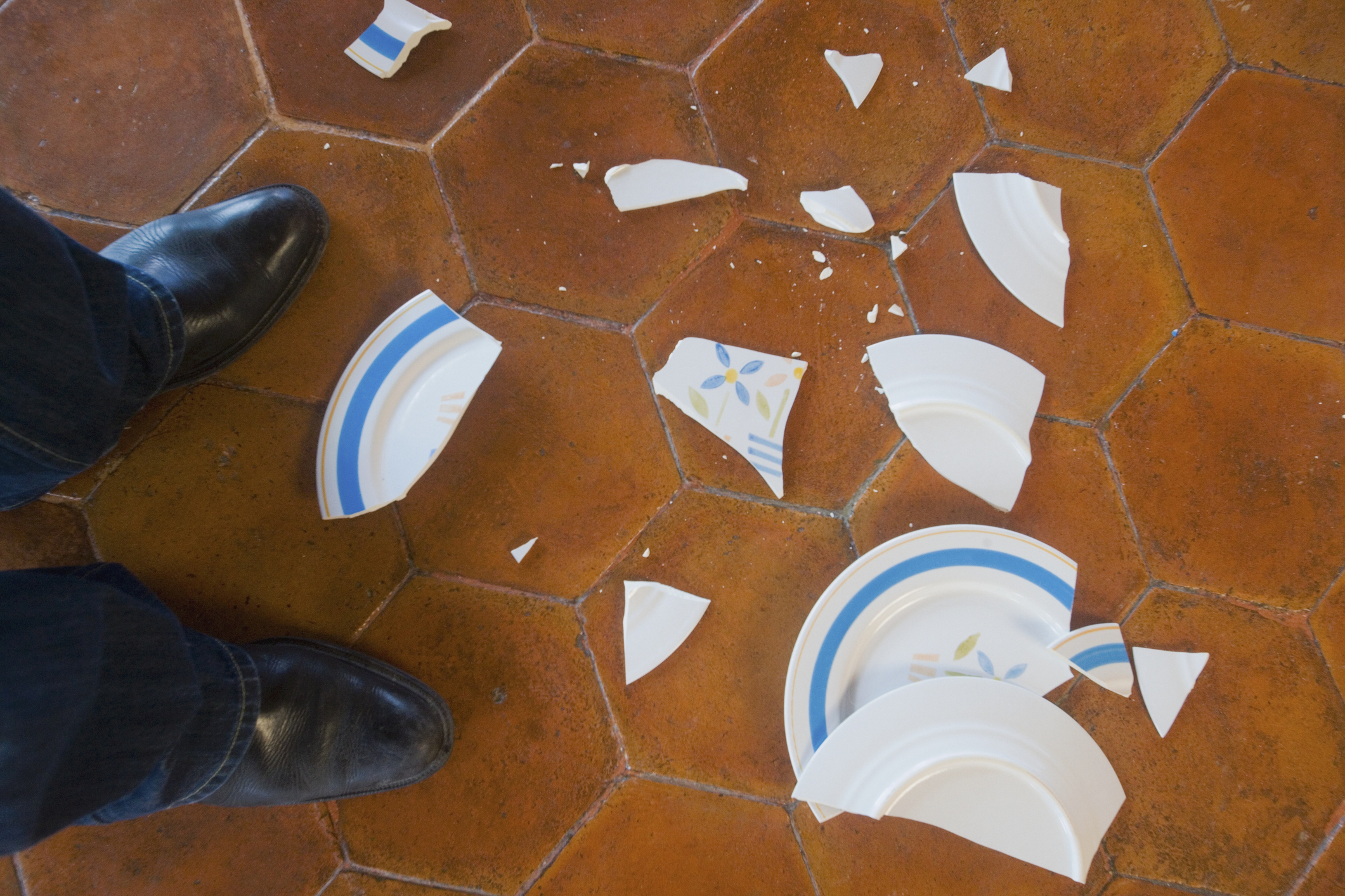 Broken white plate with blue stripes and yellow flowers scattered on the floor by two shoes