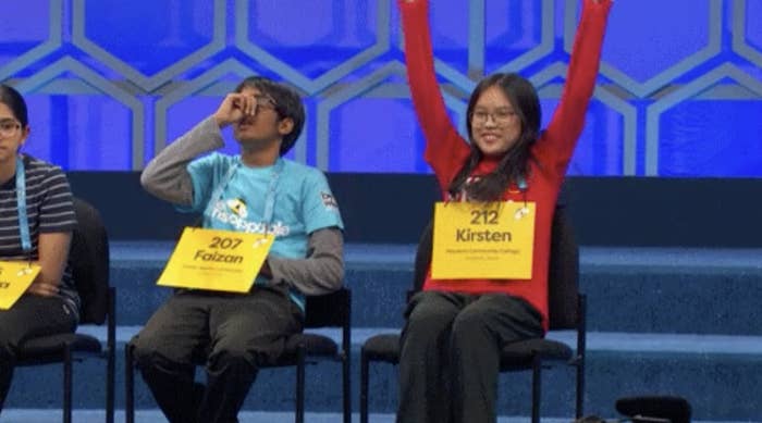 Participants Faizan (left) and Kirsten (right) at a spelling bee, with Kirsten raising her arms in celebration. Both wear numbered name tags indicating their names