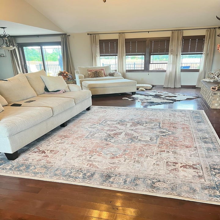 A modern living room with a large patterned rug, beige sofa, chaise lounge, and wooden floors. The room has several windows with brown blinds and curtains