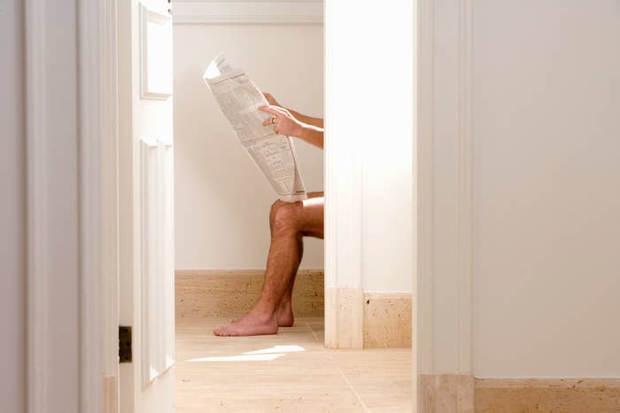 Person reading a newspaper while sitting in a bathroom, with legs and feet visible
