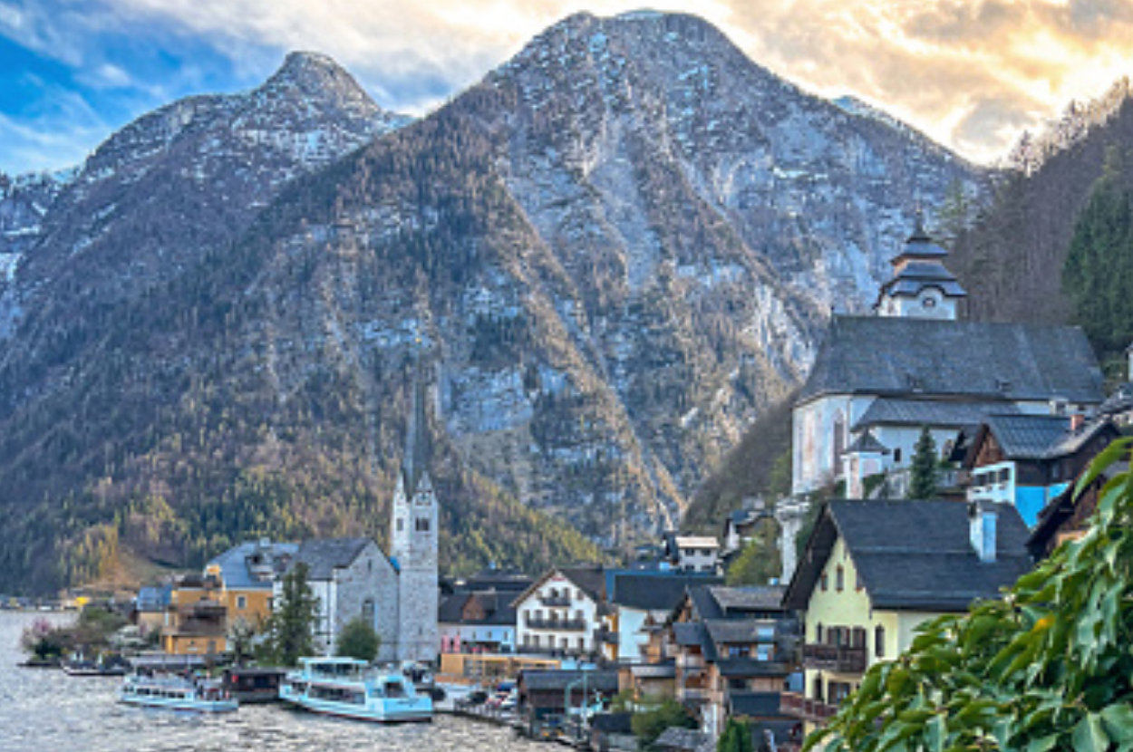 A picturesque mountain village with boats docked on a river, surrounded by towering snowy mountains. Various quaint buildings including a church are visible
