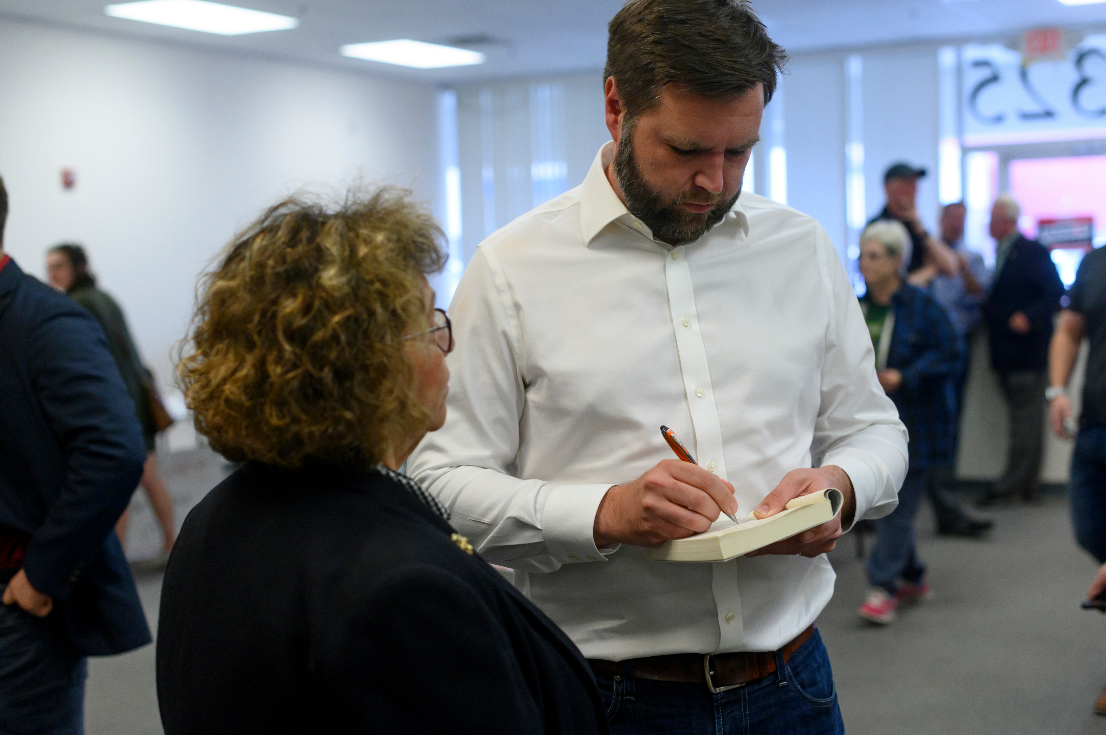 J.D. Vance signs a book while speaking with a woman at a public event attended by several people