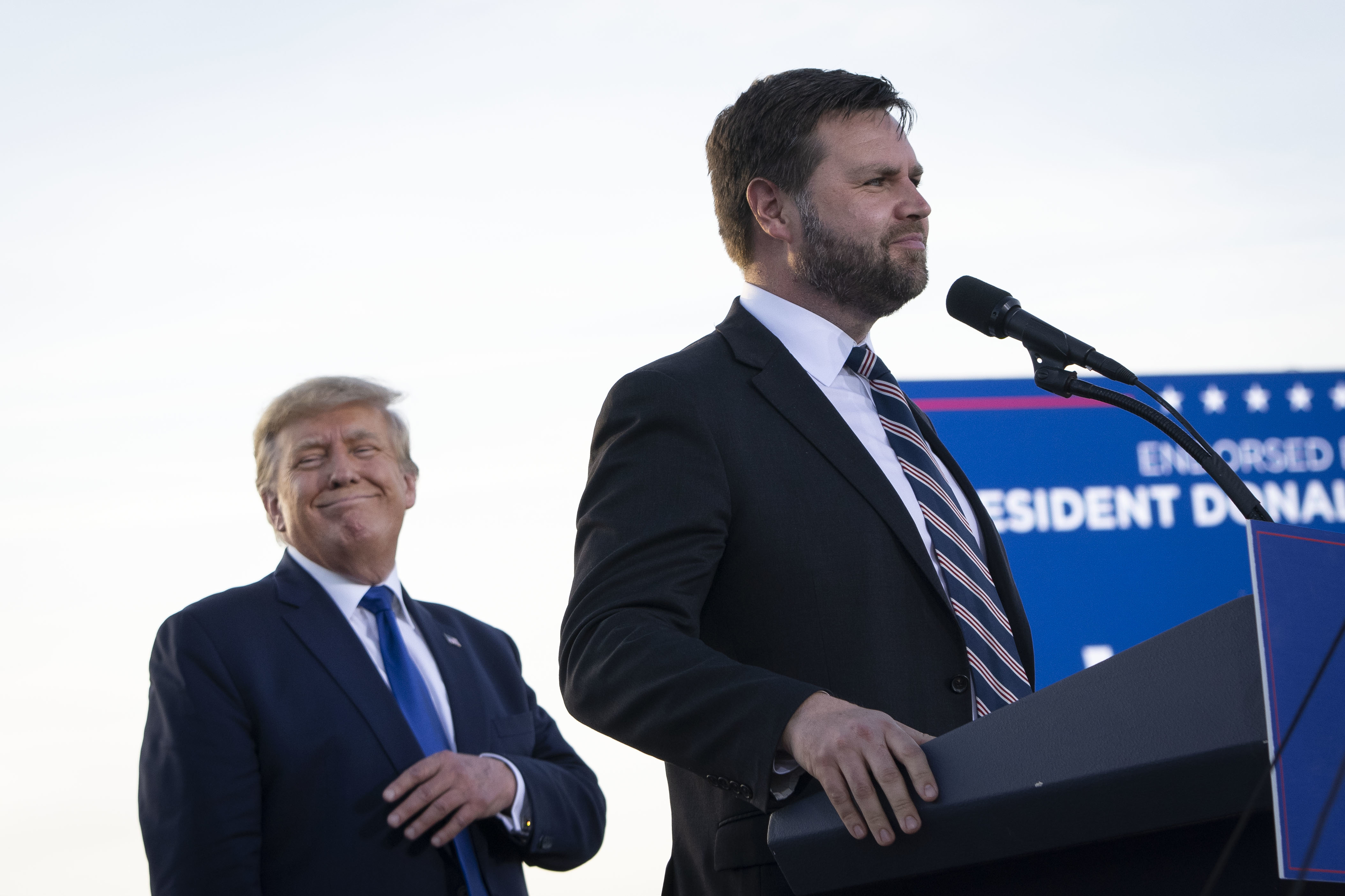 Donald Trump and J.D. Vance at a podium during an event. Trump is smiling in the background while Vance speaks at the microphone