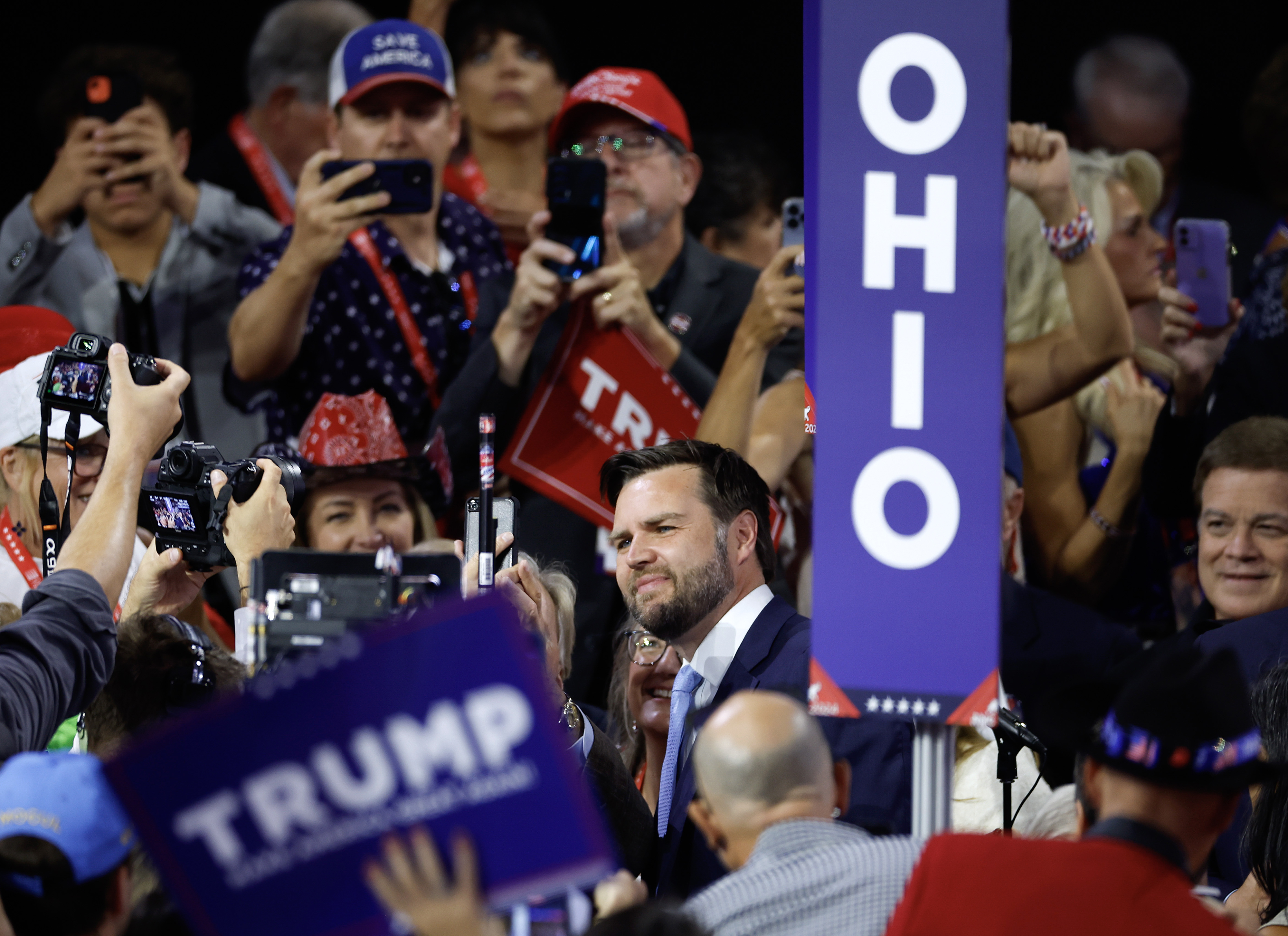 J.D. Vance at a rally with supporters holding "Trump" and "Ohio" signs, posing for photos and videos. Crowd surrounds him, capturing the moment on their devices