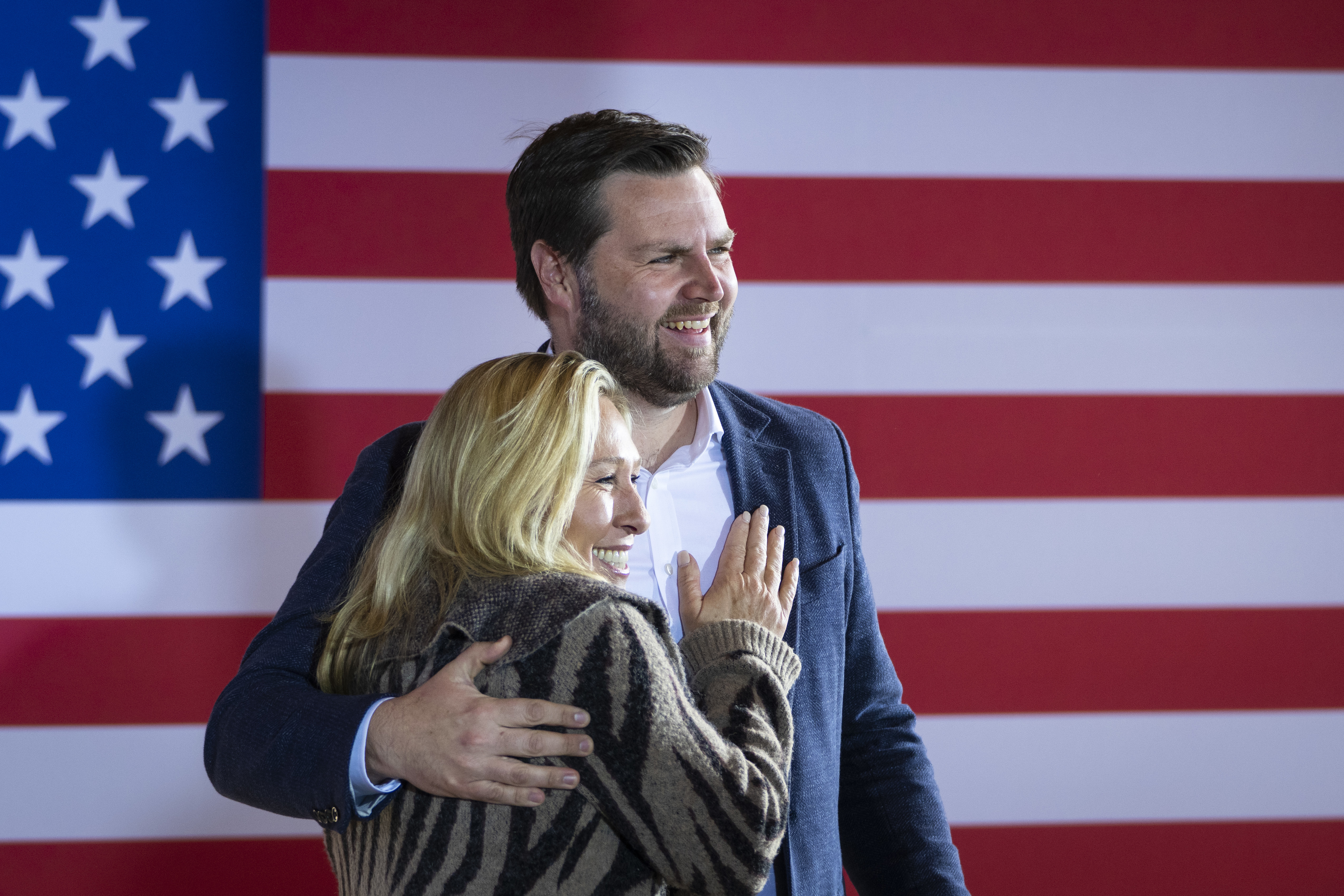 Marjorie Taylor Greene and JD Vance stand together, smiling, in front of an American flag backdrop