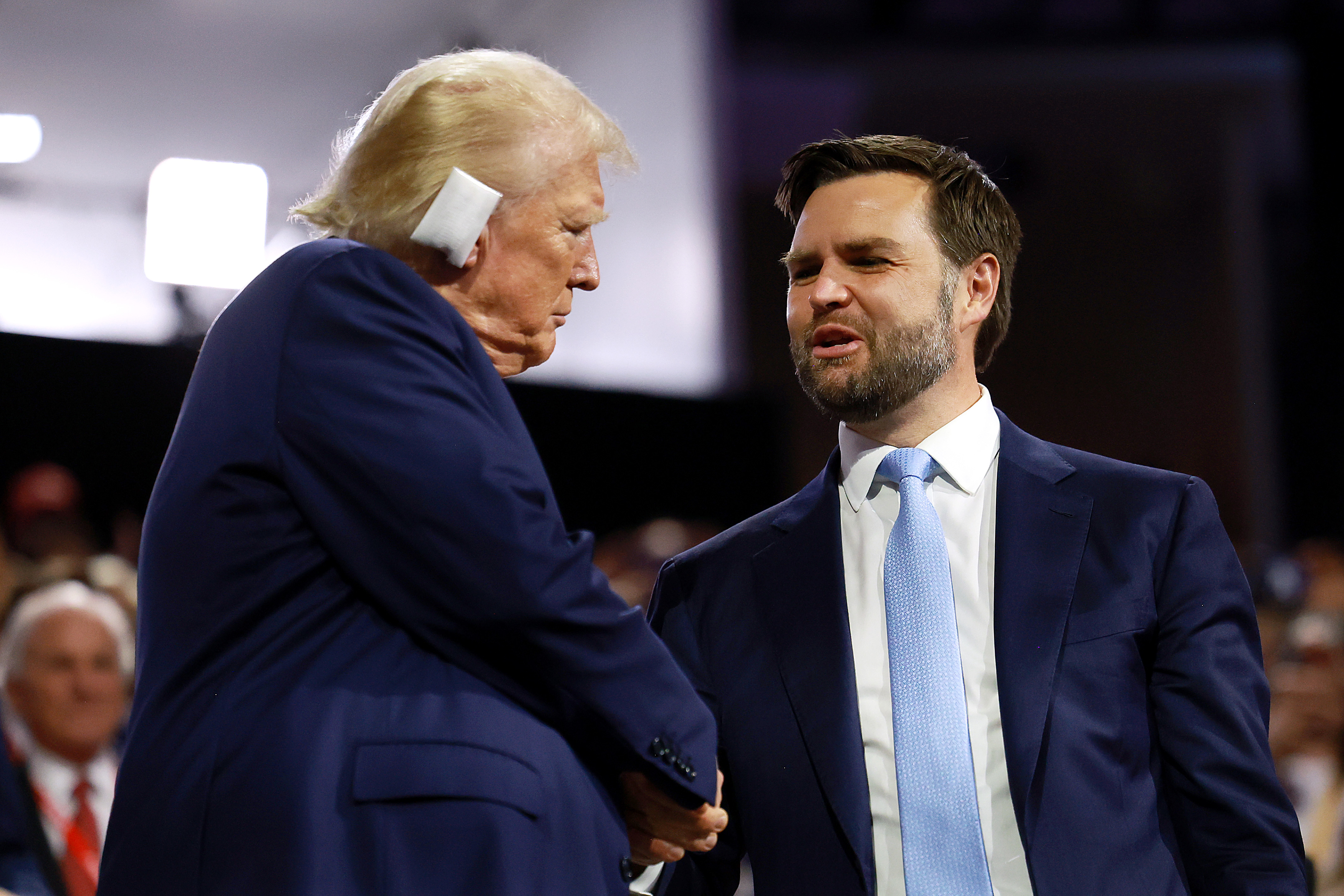 Donald Trump and JD Vance, both in suits, are shaking hands and conversing at an indoor event