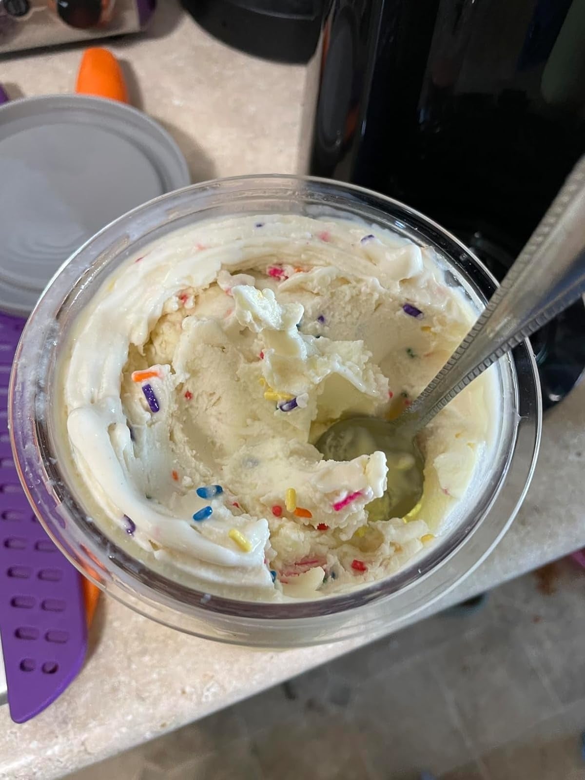 Partially eaten instrumentality  of creamy frosting with colorful sprinkles and a metallic  spoon inside. Lid and a purple utensil nearby