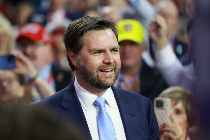J.D. Vance at a public event in a suit and tie, surrounded by a crowd who appear to be cheering and taking photos