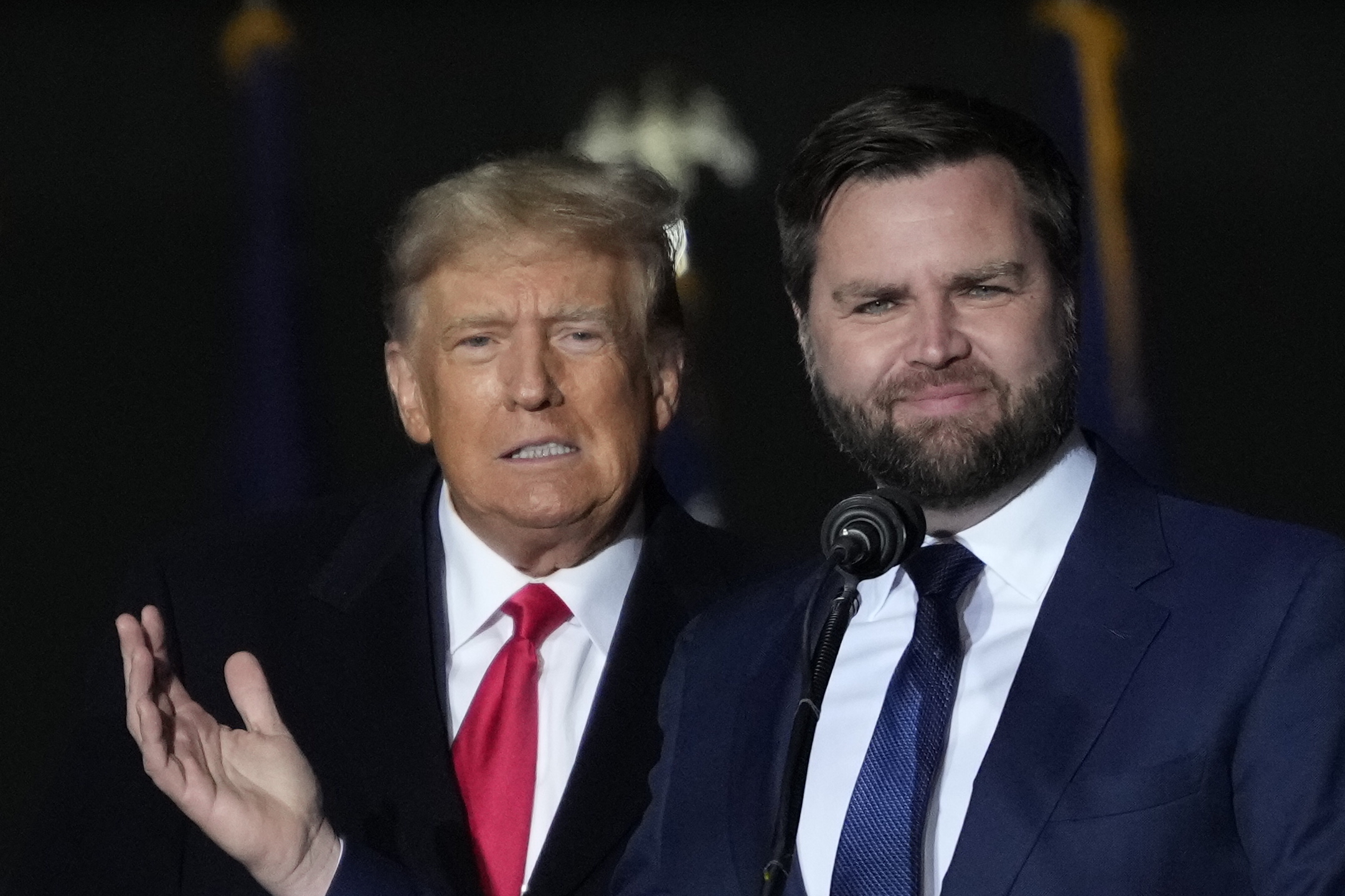 Donald Trump in a dark suit and red tie stands next to J.D. Vance in a dark suit and blue tie, speaking at an event