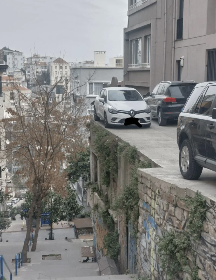 White car precariously parked on the edge of a building, overlooking a city street. Several other cars and buildings are visible in the background