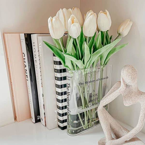 Bookshelf with books, white tulips in a vase labeled &quot;The Mystery of Growth,&quot; and a modern sculpture of a seated person