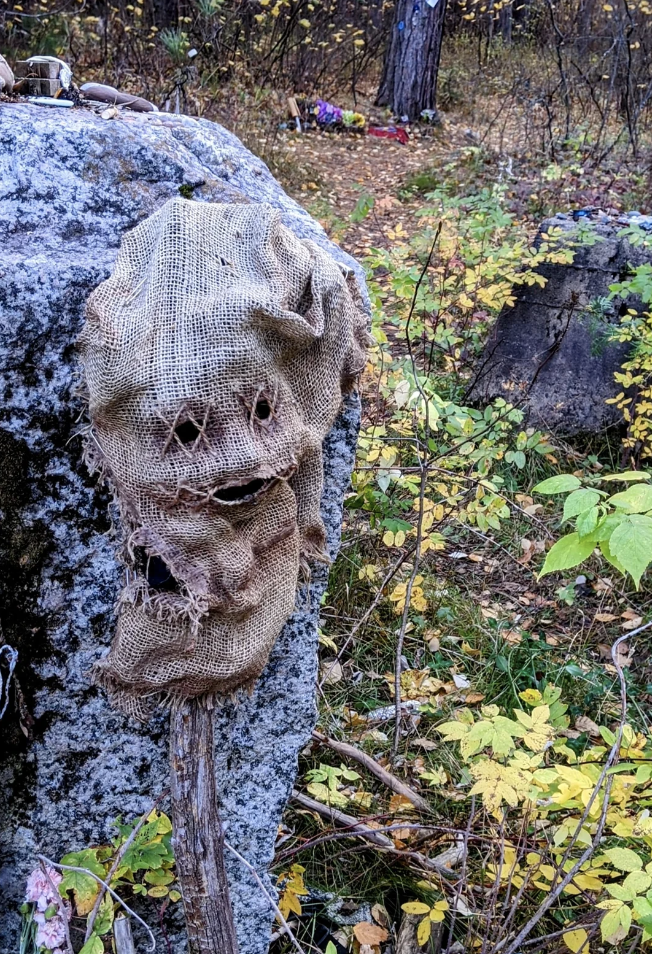 A burlap sack with holes resembling a face is draped over a stick, placed against a rock in a forested area