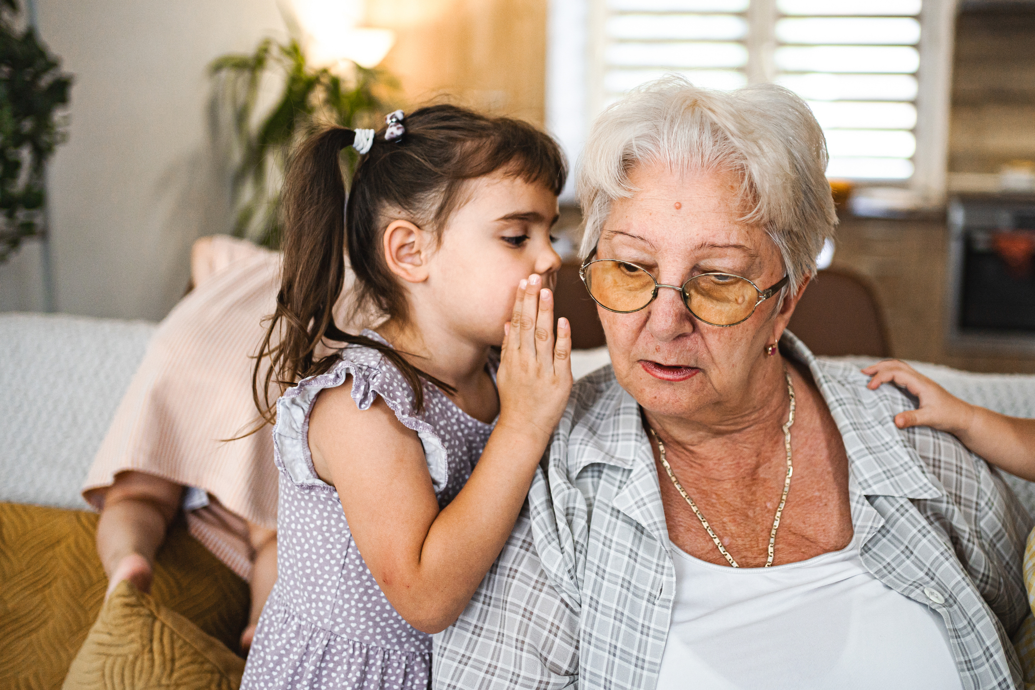 A young girl whispers into her grandmother's ear as they sit on a couch. The grandmother, wearing glasses and a casual outfit, listens attentively