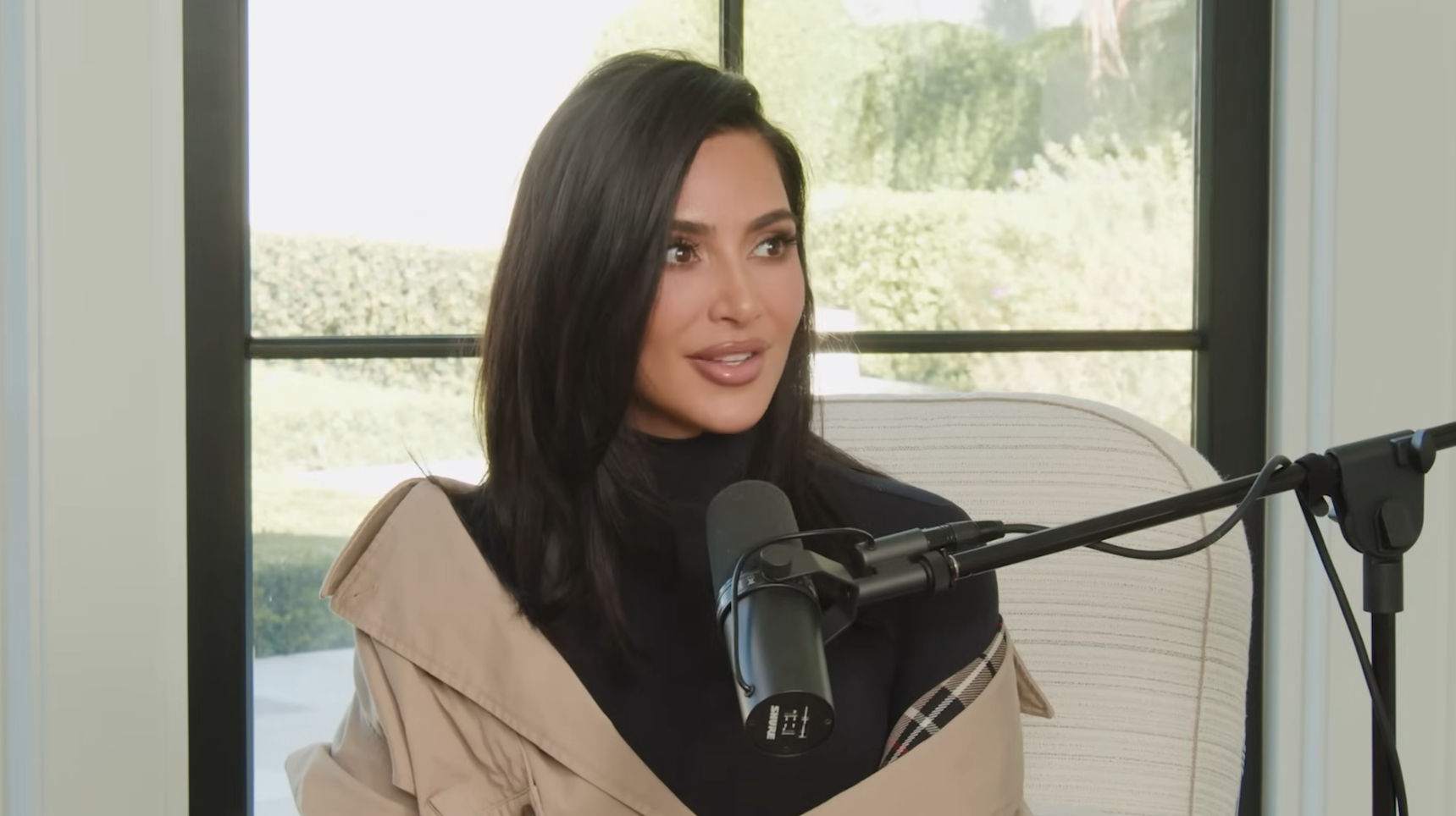 Kim Kardashian is seated at a microphone during an interview, wearing a stylish trench coat over a black turtleneck